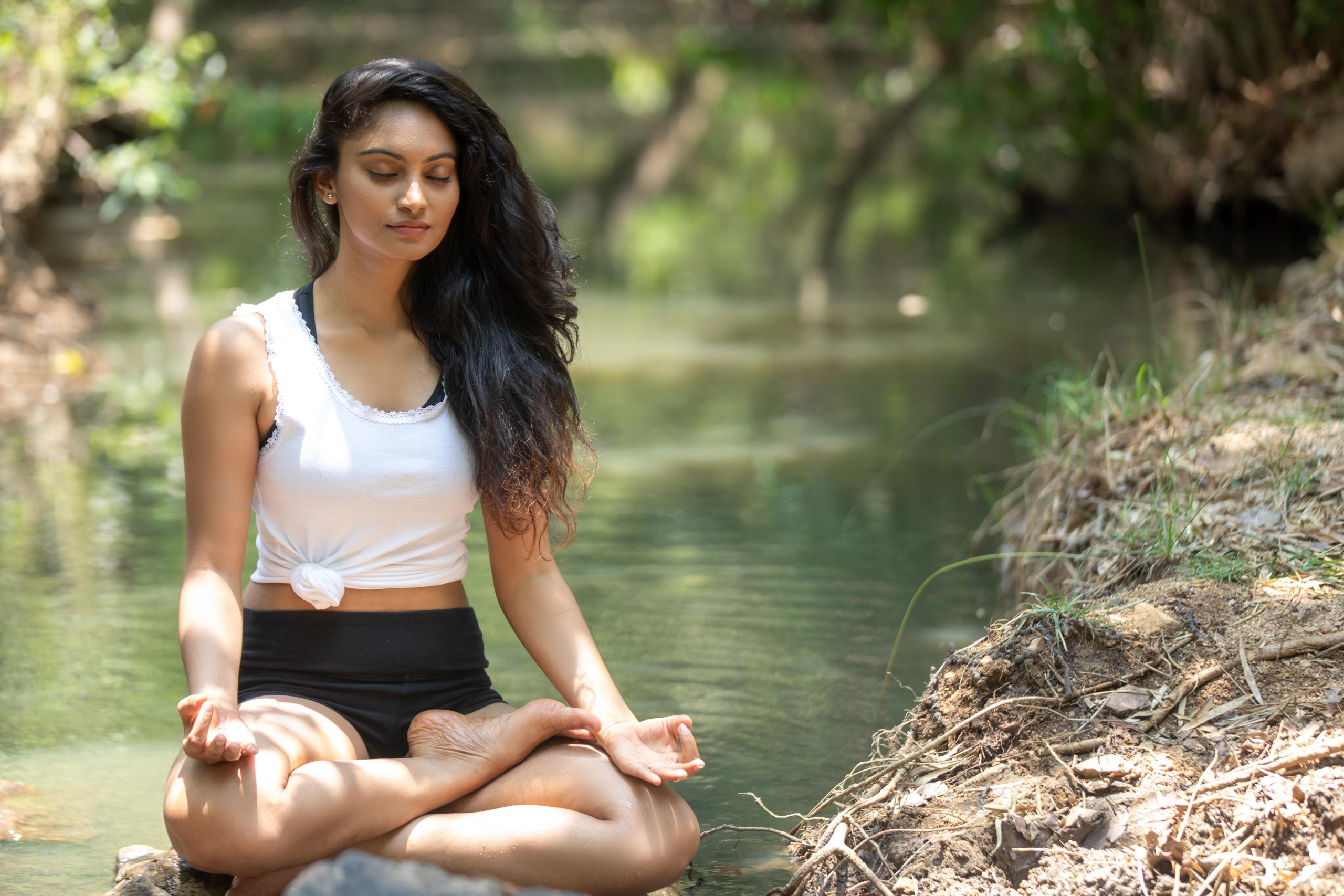 Woman meditating beside a calm river in nature, embodying inner peace and mindful living